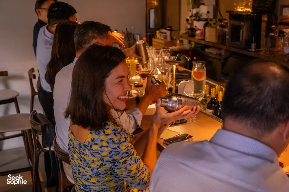 Small group gathered at the bar counter in Singapore, watching a mixologist prepare cocktails amid bottles and glassware.