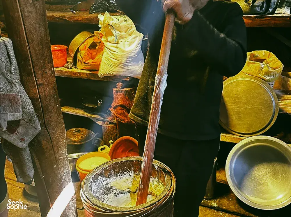 During a Khampa Caravan homestay meal visit in Shangri-La, an apron-clad cook stirs a pot beside simmering utensils