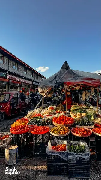 Lijiang Old Town market street with produce and snack stalls under tarpaulins beside parked scooters at dusk