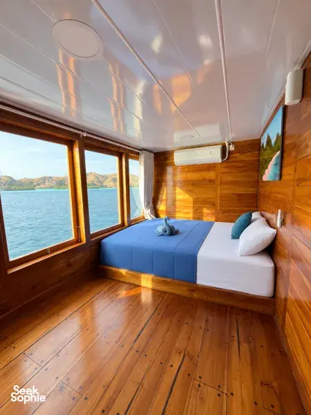 Wooden phinisi cabin with two beds and sunset light through panoramic windows on a shared Komodo cruise from Labuan Bajo