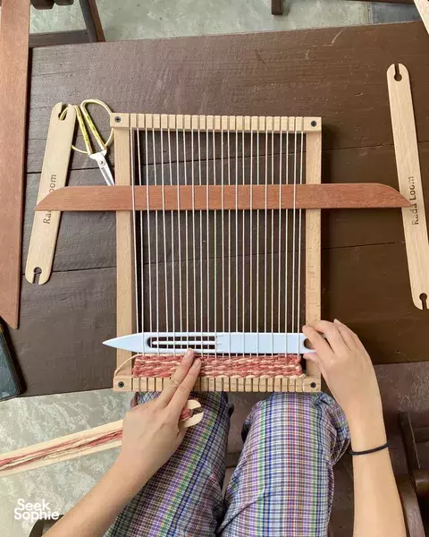 Hands weaving on a wooden Thai loom at Kalm Village weaving workshop, Chiang Mai, with colourful threads and heddles