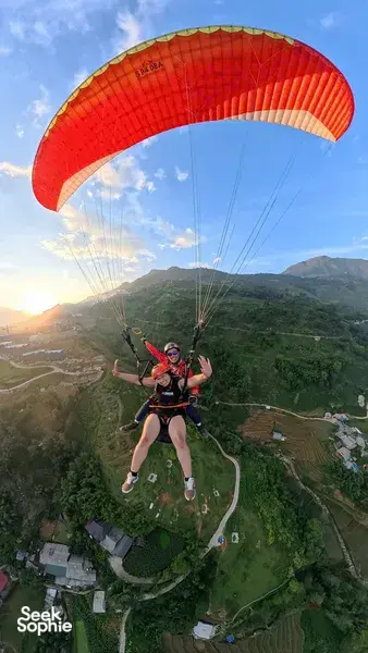 A paraglider duo flies beneath a bright red canopy over winding roads and villages during golden hour in Sapa