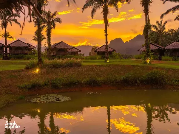 Palm-lined campground in Thailand at sunset, with warm sky reflections over a pond and thatched-roof villas