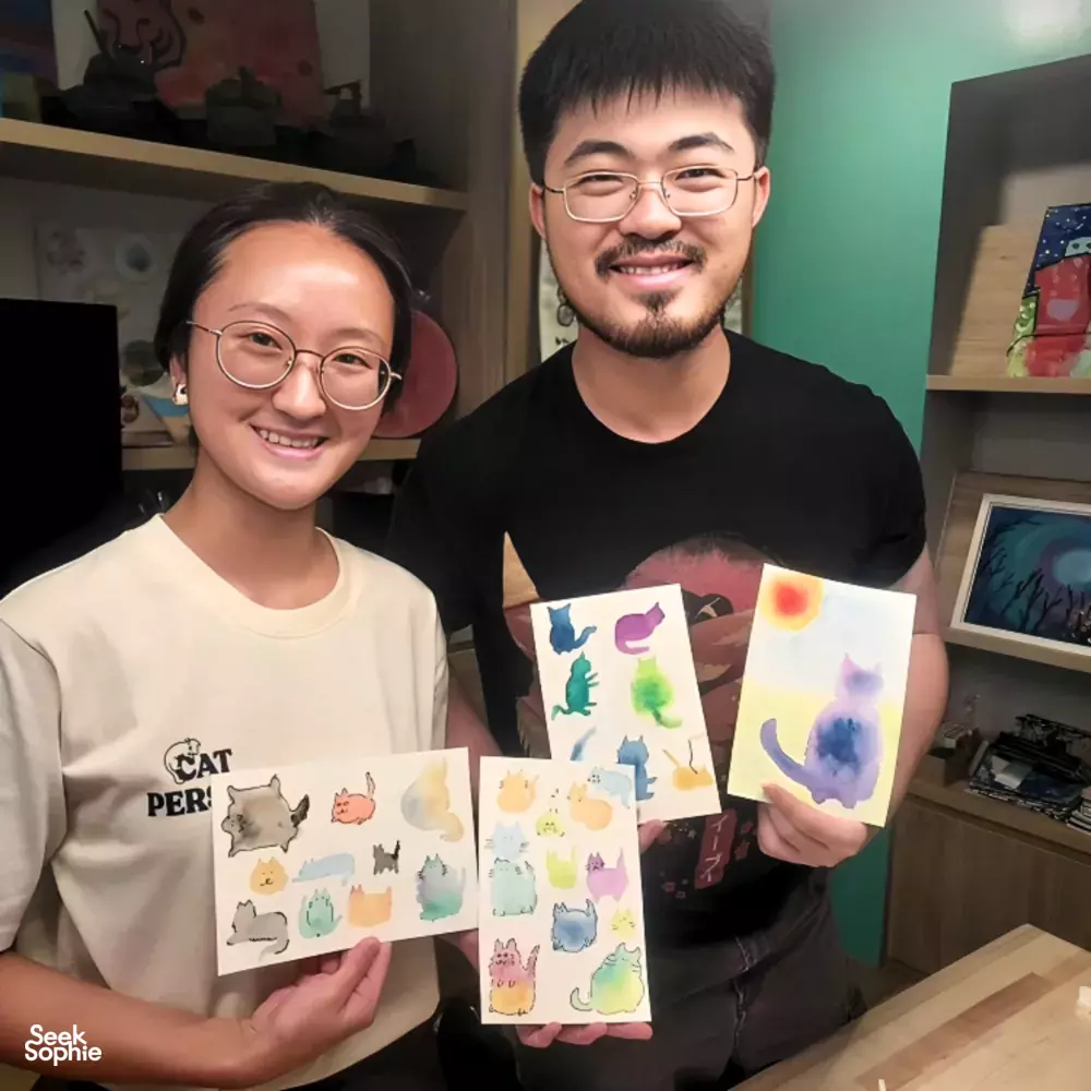 Art For Good art therapy in Singapore: two participants smiling while holding cat-themed paintings during workshops in Singapore
