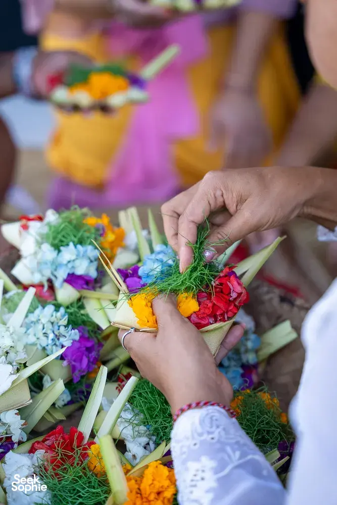 Hands building canang sari leaf baskets with colourful flowers on woven trays during a ceremony in Ubud