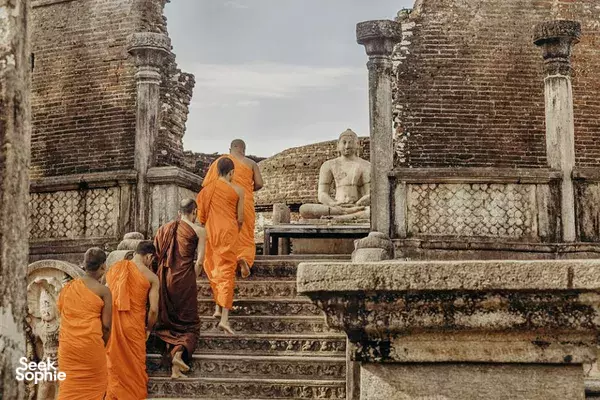 Orange-robed Buddhist monks walking past a seated Buddha statue among ancient stone ruins in Polonnaruwa, Sri Lanka
