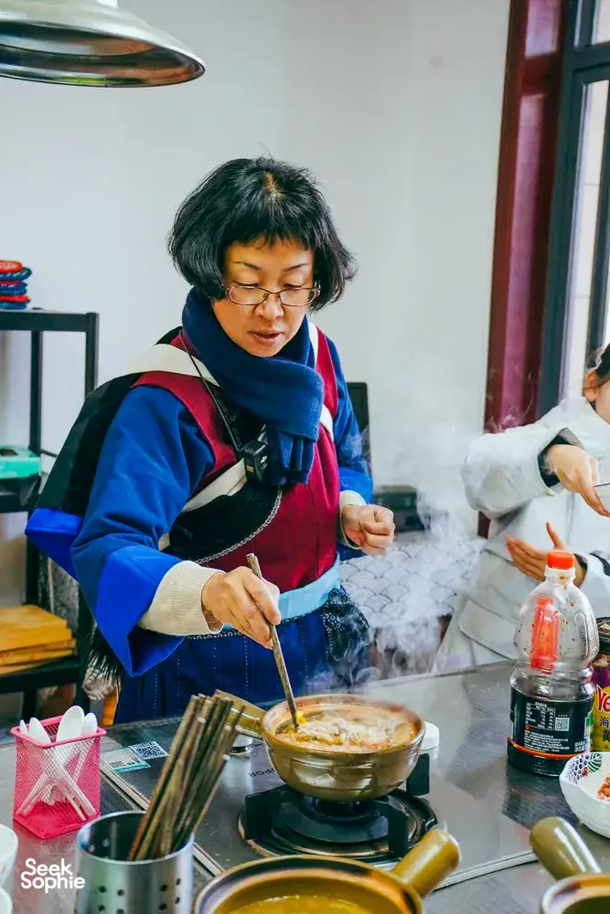 Fancy stirring a pot on a stovetop while cooking Yunnan food in a Lijiang kitchen, with herbs and sauces on the table