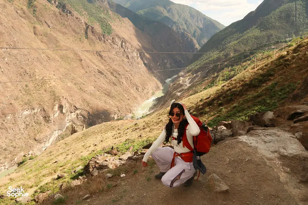 Hiker pausing on a rocky viewpoint along the high trail of Tiger Leaping Gorge near Lijiang, with a vast canyon below