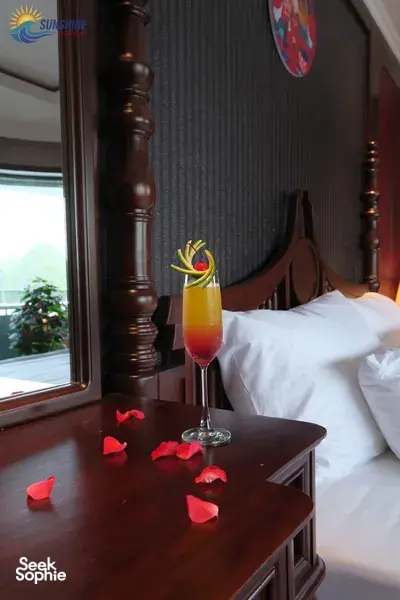 Cocktail with a flower garnish on a bedside table beside rose petals in a luxe Cat Ba cruise cabin by Sunshine Golden Cruises