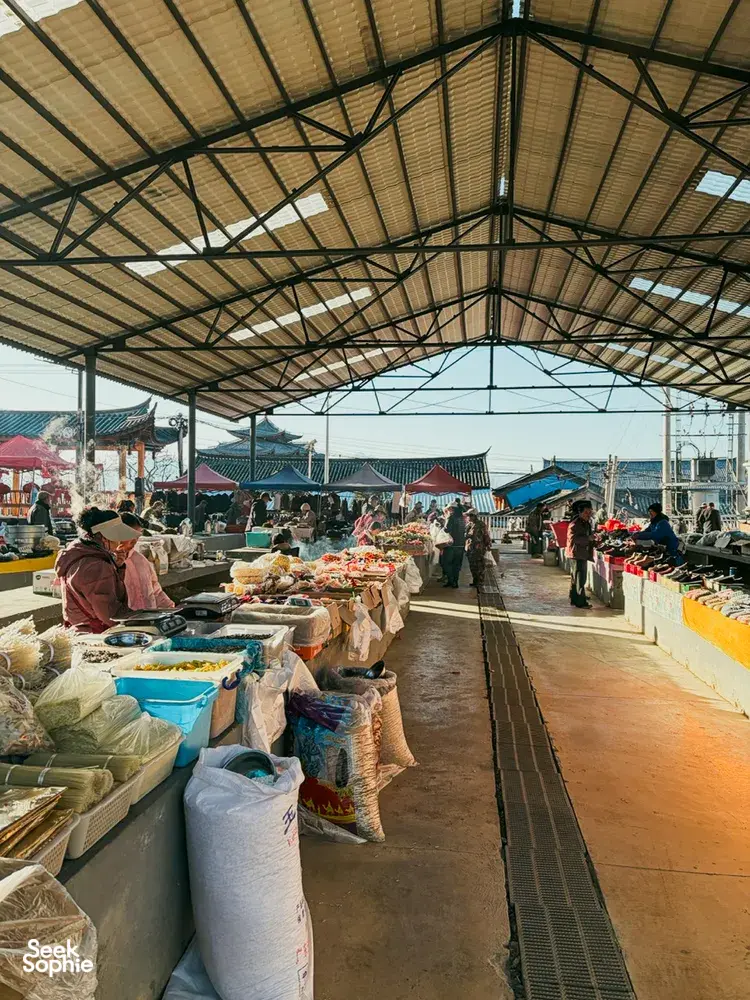 Long covered village market lane with stall tables of packaged goods and fruit, during a guesthouse stay trek in Lijiang, Yunnan