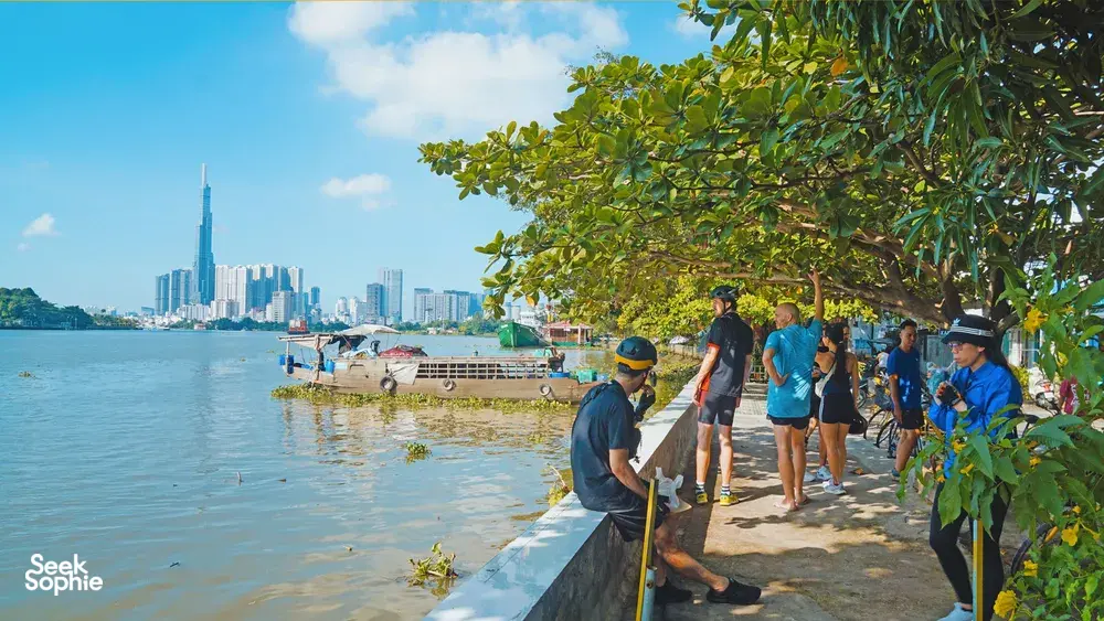People walking along a quiet riverbank path on Mekong Island, with Ho Chi Minh City skyline across the water