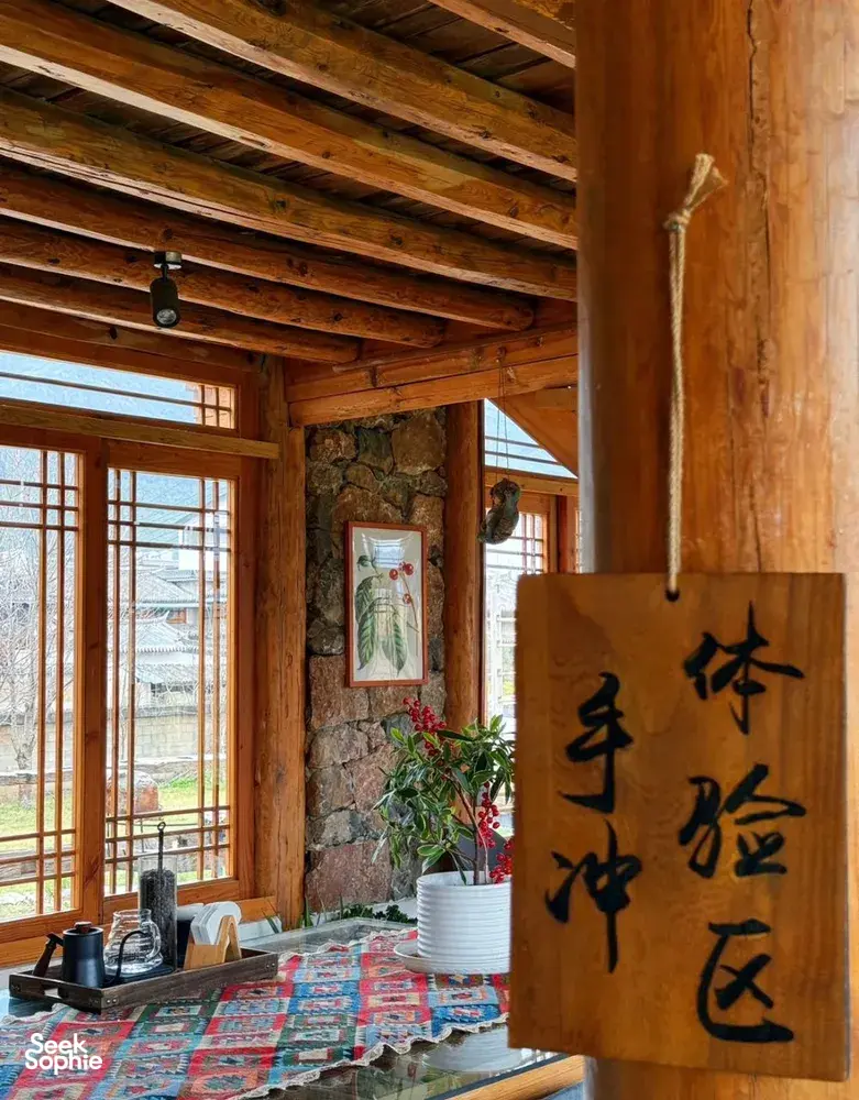 Rustic wooden tea room interior with large pinned calligraphy panel, stone wall and windows, with cups and kettle on a table in Lijiang