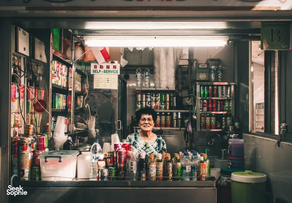 Chef in a Singapore hawker stall serving from behind a counter lined with jars and tins, with a “self-service” sign