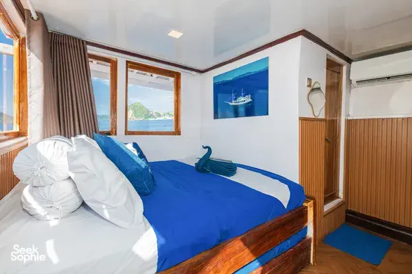 Cabin bedroom on a Komodo boat trip in Labuan Bajo, with blue bed linen, ocean-view windows, and folded bedding