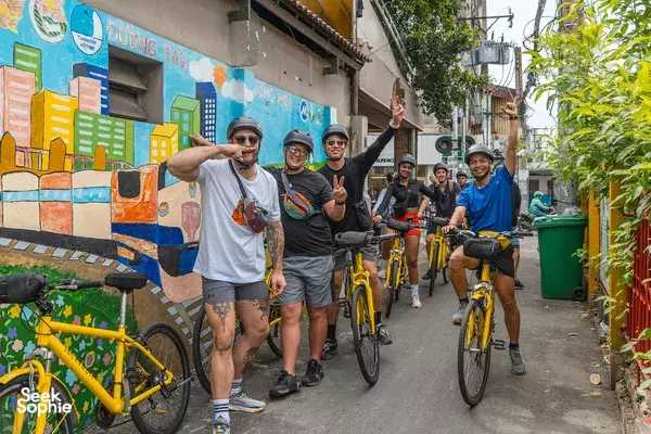 Group of cyclists smiling in a narrow alley beside a colourful mural wall in Ho Chi Minh City during a bike tour