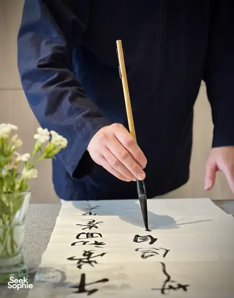 Hand gripping a calligraphy brush writing bold Chinese characters on rice paper in a Singapore workshop