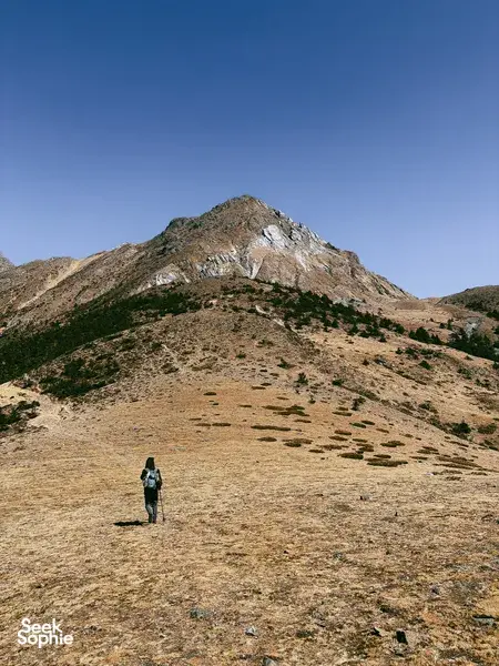 Solo hiker crossing a barren alpine slope toward a jagged peak on a guided hike of Jade Dragon Snow Mountain in Lijiang