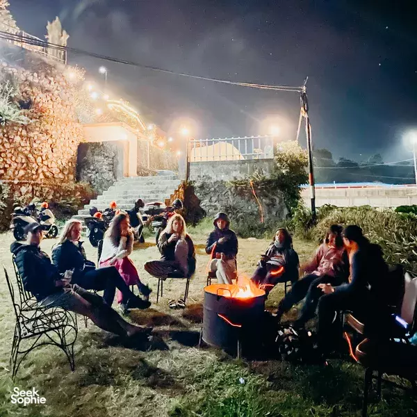 Travellers gather around a glowing fire pit at night in a Lombok village, with scooters parked along the roadside