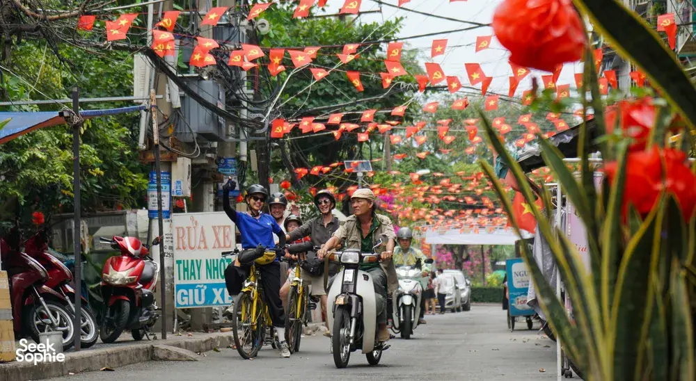 Cyclists and motorbikes cross a decorated Vietnamese street under red bunting flags during a Mekong Island bike tour