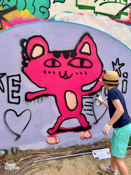 Painter adding details to a pink cartoon cat mural on a wall, with spray cans and paint buckets during a creative workshop in Da Nang