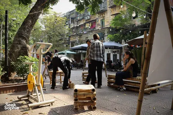 Perhimpunan komuniti di halaman redup di Hanoi, dengan orang ramai bersantai berhampiran tempat duduk luar dan buaian di bawah pokok.