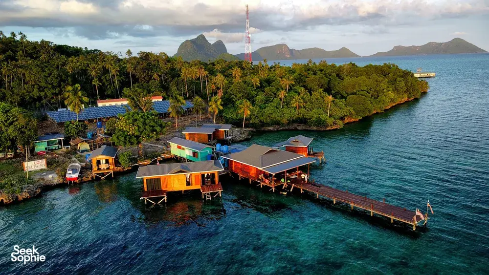Aerial view of Semporna sea lodge cabins along the shoreline with a long jetty, boats and distant hills under evening light