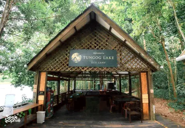 Open-air wooden pavilion with a sign reading Welcome to TUNGOG LAKE ECO CAMP in a leafy rainforest setting