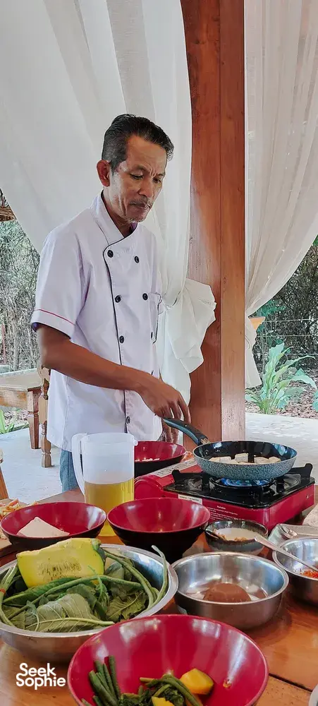 Chef Michael stirs on a skillet beside red bowls and plates during a Traditional Manggarai vegan cooking class in Labuan Bajo