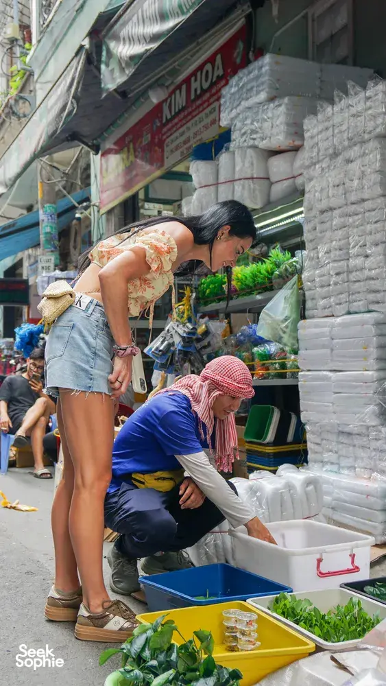 一位穿着花上衣的旅行者在胡志明市参加自行车之旅时,浏览街头市集摊位,旁边是装满新鲜香草的托盘