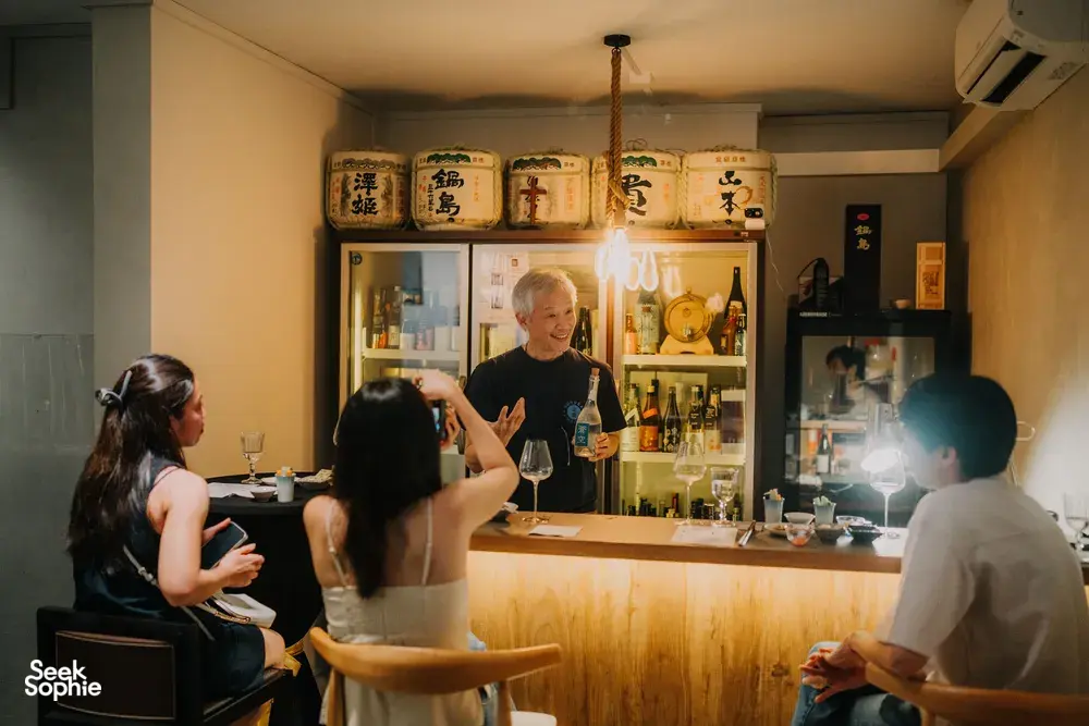 Host pouring rare sake for guests at a small bar counter, with labelled bottles and lantern light in Singapore — Singapore hidden gems