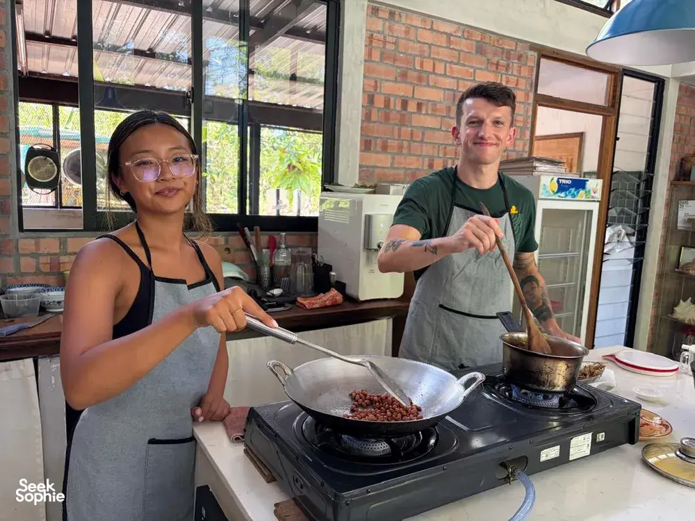 Woman and man cooking at a stovetop in Kebun Lidstari’s Langkawi farmhouse kitchen, preparing ingredients on a frying pan