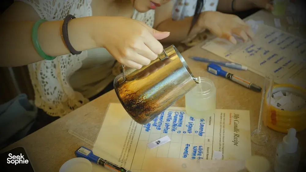 Hand pouring wax into a small candle jar on a Singapore worktable, with fragrance bottles and labels beside it in a perfume workshop