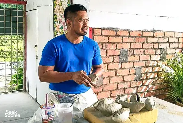 Ceramic artist in Ilham Ceramic Studio, Langkawi, shaping pottery at a table outdoors beside a brick wall