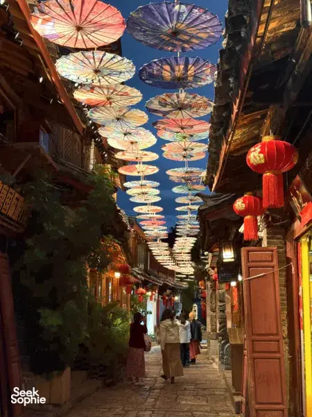 Lantern-lit lane in Lijiang’s old town with colourful paper umbrellas overhead as travellers stroll at dusk
