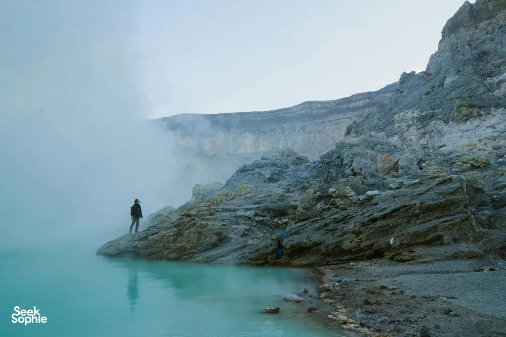 Hiker standing on a foggy ledge beside a turquoise lagoon near jagged cliffs, with distant clouds in the valley