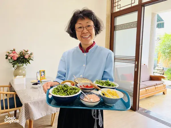 Smiling chef in a blue jacket serving multiple bowls of Chinese dishes on a dining table beside an open window in China