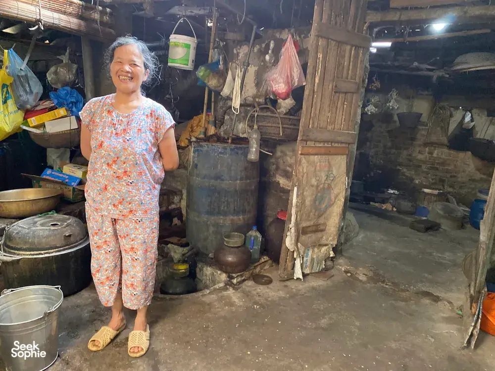 Smiling woman standing in a rustic kitchen with cooking pots during a traditional tofu-making visit in Hanoi’s Red River village
