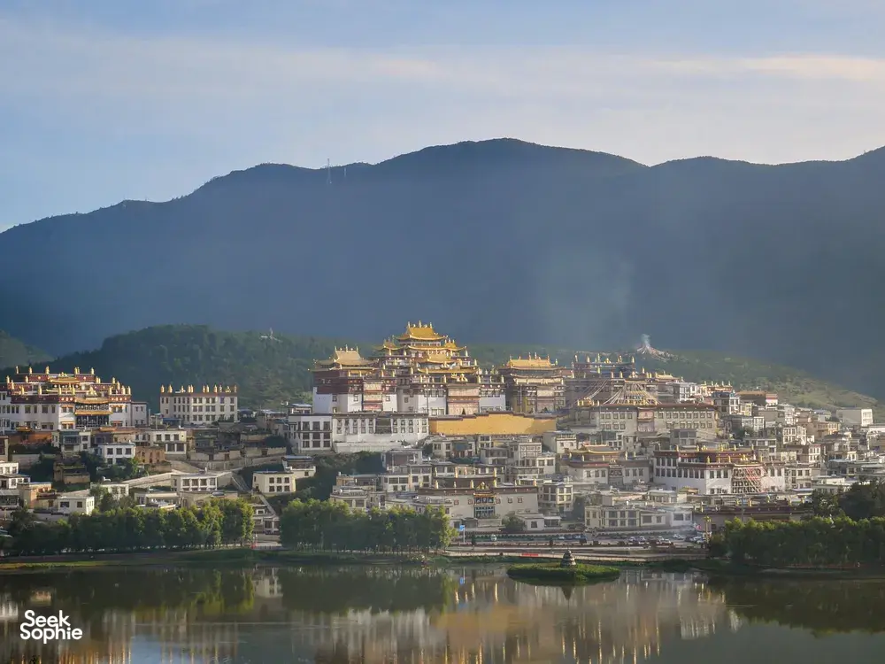 Riverside panorama of a hillside Chinese town with a gold pagoda, layered buildings, and misty mountains reflected in the water
