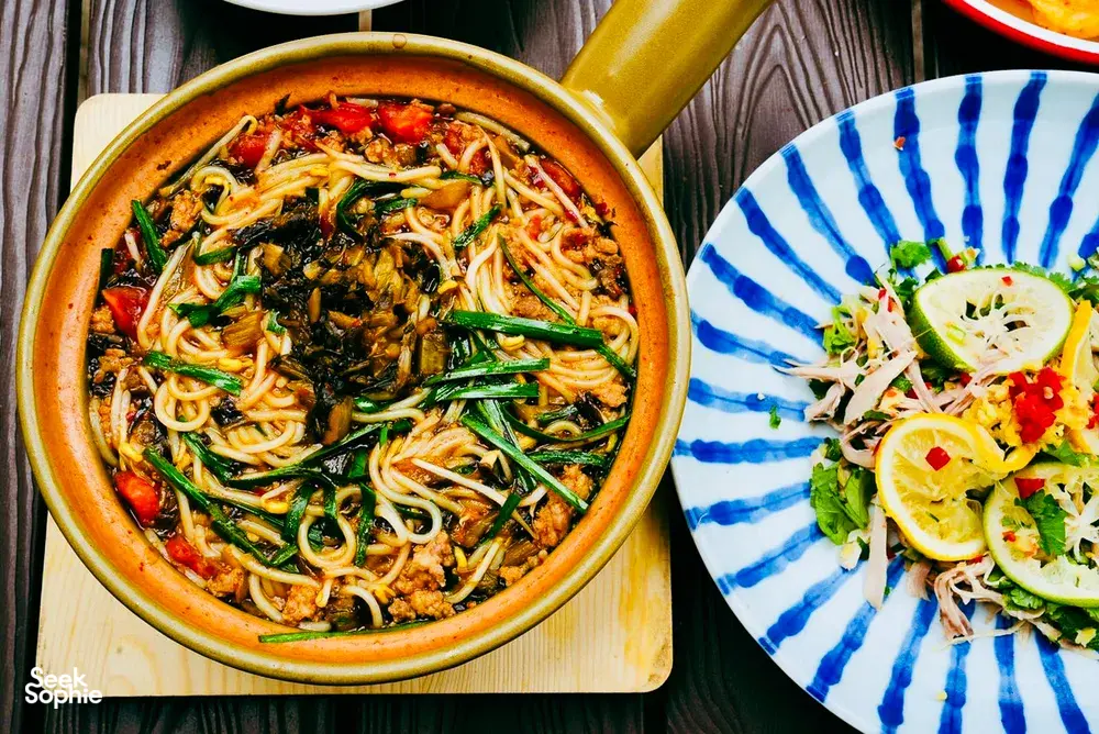 Steaming Yunnan-style noodle broth with greens and red chillies in a terracotta pot, plus a lemony side salad in Lijiang