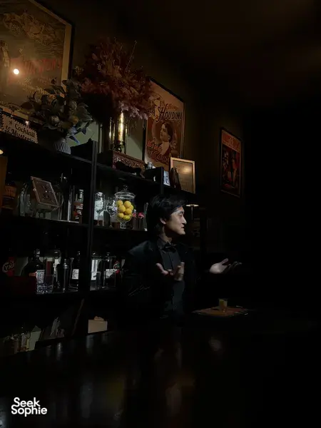 Performer gesturing from behind a bar counter at The Magic Bar in Singapore, dim lighting and glass bottles around