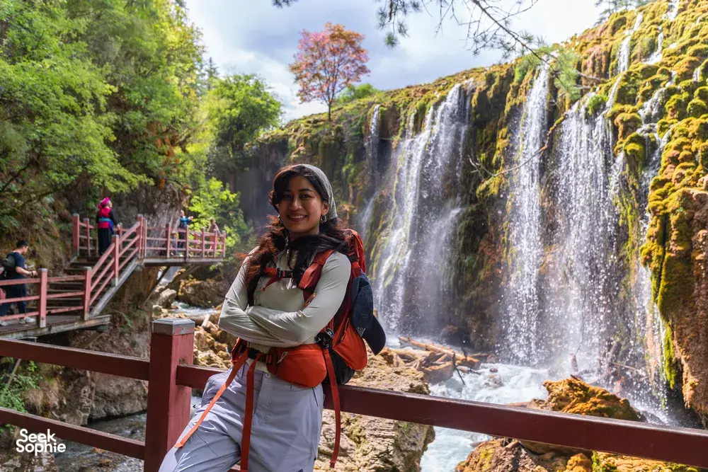 Hiker posing by a multi-tier waterfall in Shangri-La, standing on a wooden platform with red handrails and trekking gear