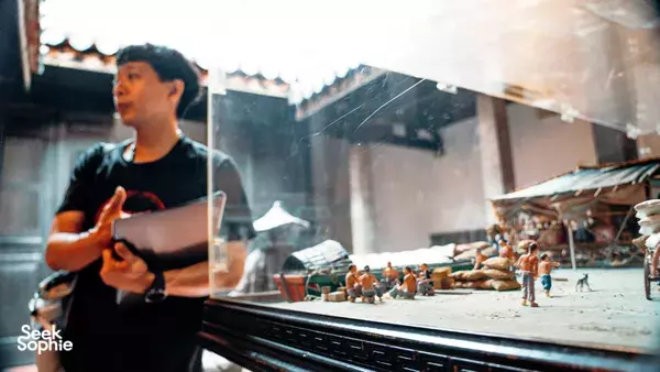 Triad Trails guide speaking beside a glass-fronted street market diorama with stalls and miniature shoppers in Singapore