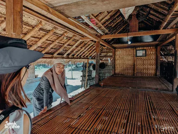 People working at a long wooden slat loom inside a traditional Lombok village house, with one person leaning over the slats