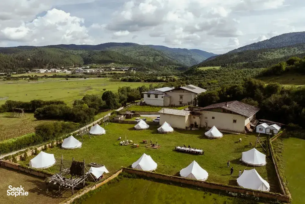 Aerial view of a Tibetan mountain valley camp at Ferme Liotard in Shangri-La with white tents, paths and surrounding hills