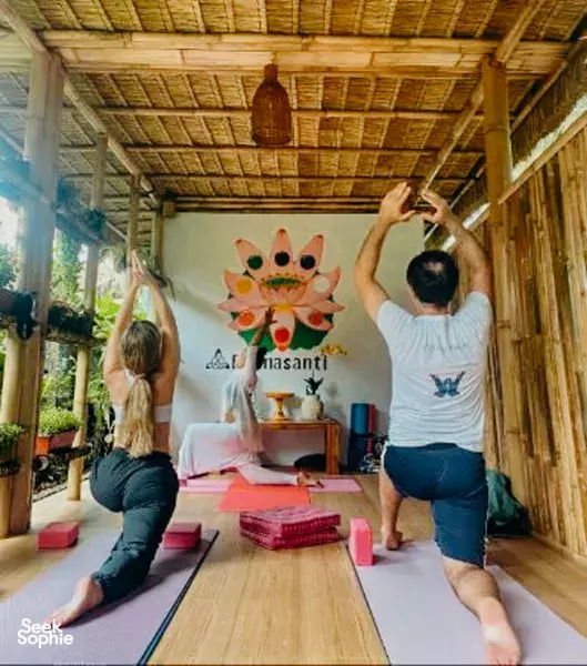 Yoga class in a thatched-roof bamboo studio at Pranasanti Yoga, participants raising arms on mats during a group session in Ubud