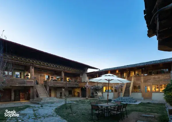 Ferme Liotard courtyard with traditional wooden buildings, evening lights, and outdoor dining chairs beside a white umbrella
