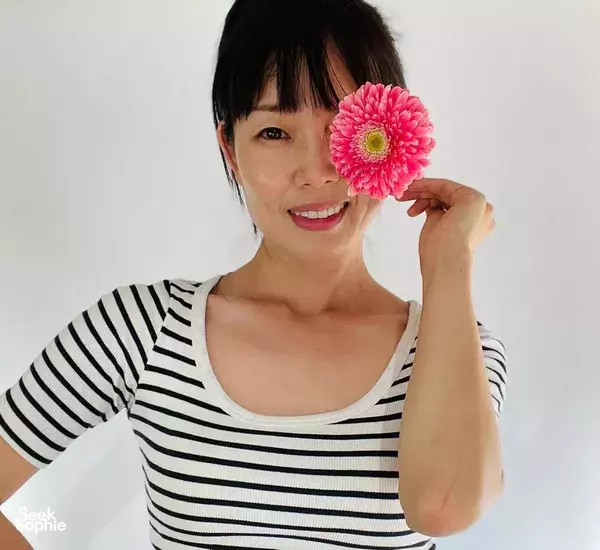 Smiling woman holding a single bright pink flower in front of her face against a light background, in Singapore