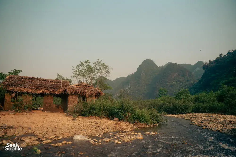 Thatched-roof stilt hut by a rocky stream with hazy limestone mountains in Phong Nha on a local-tribe tour