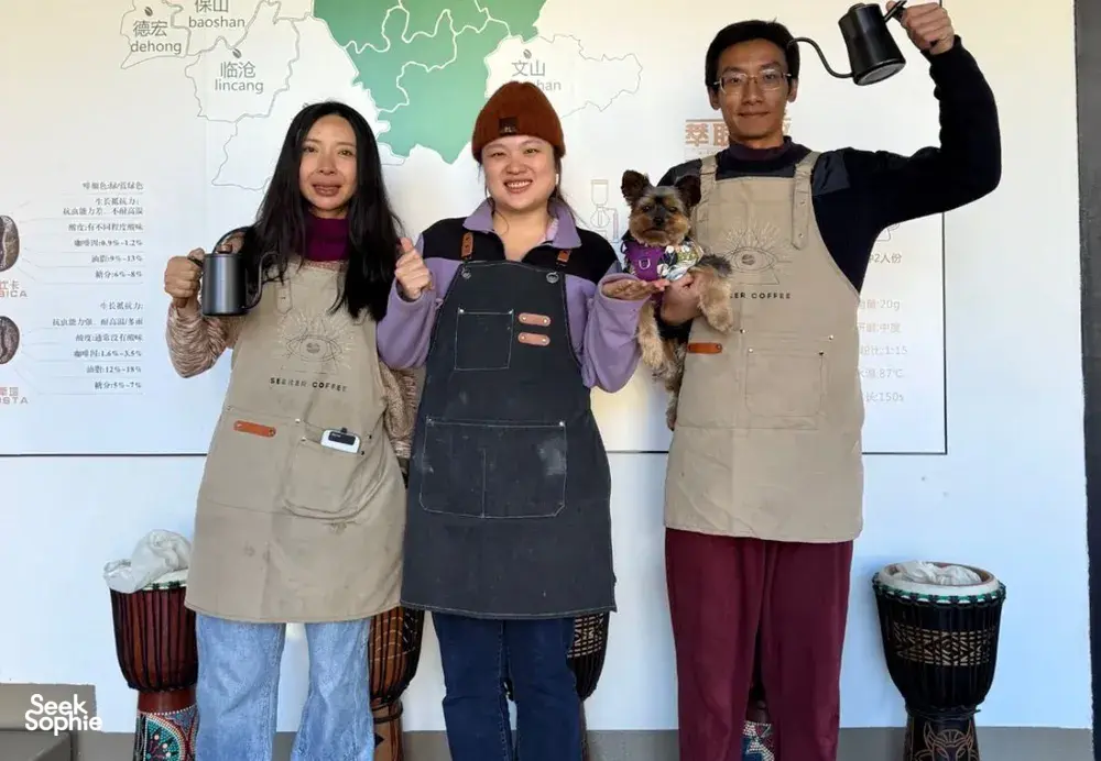 Guests in aprons posing with a small dog in front of a Yunnan coffee map at a coffee workshop in Lijiang
