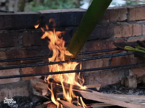 Ngapuh Homestead campfire cooking over open brick hearth in Kuching’s rainforest, with bamboo pole and flame visible